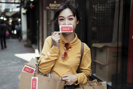 美丽的亚洲女人穿着黄色毛衣，背着许多贴着价签的购物袋，走在街上。