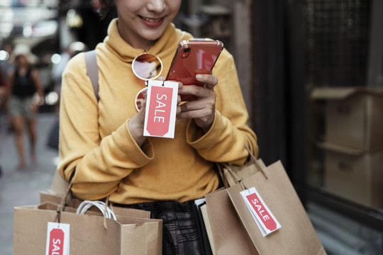 开朗的亚洲女人用智能手机上网购物，拎着购物纸袋，走在街上。
