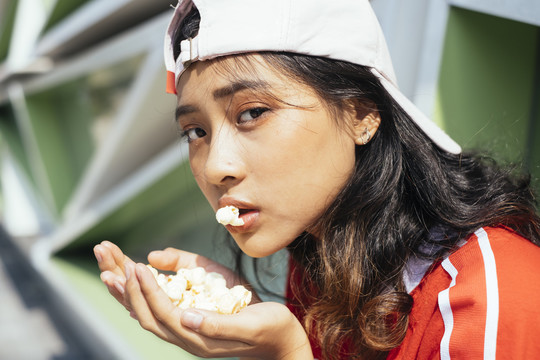 特写-年轻迷人的亚泰裔女子黑发红衫戴帽子在户外吃爆米花。