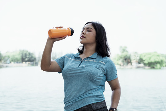 一个胖乎乎的亚洲女人穿着蓝色运动服，在大自然的湖边喝着橙色瓶子里的水。