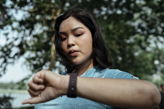 胖乎乎的运动型亚洲女性，穿着蓝色运动服，在公园慢跑后在智能手表上查看时间。