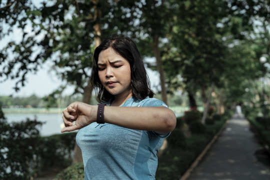 在公园慢跑后，一位穿着蓝色运动服、身材胖胖的亚洲女士在智能手表上查看时间。