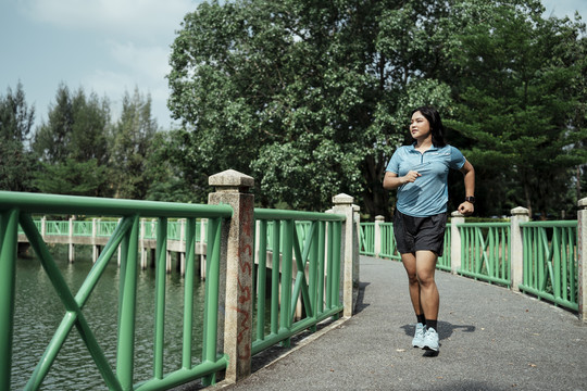 胖乎乎的运动型亚洲女人穿着蓝色运动服在湖边的自然公园跑道上跑步。