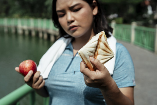 艰难的选择-胖乎乎的运动型亚洲女人穿着蓝色运动服选择苹果和三明治。健康的或不健康的。