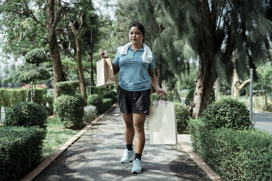 胖乎乎的运动型亚洲女人穿着蓝色运动服去购物，在公园运动后拿着购物纸袋。