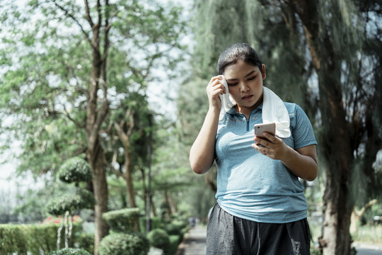 疲惫胖胖的运动型亚洲女性，穿着蓝色运动服，满头大汗，白毛巾，看着智能手机，查看网站，和朋友聊天。