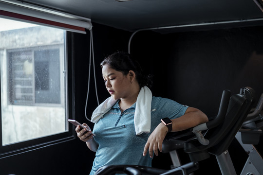 胖乎乎的运动型亚洲女性，穿着蓝色运动服，厌倦了在健身房里用智能手机锻炼。