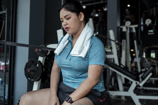 穿着蓝色运动服的疲惫胖乎乎的运动型亚洲女人在健身房辛苦锻炼后，在长凳上放松。