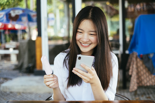 漂亮的深色长发年轻大学生穿着校服用智能手机和吃冰激凌棒。