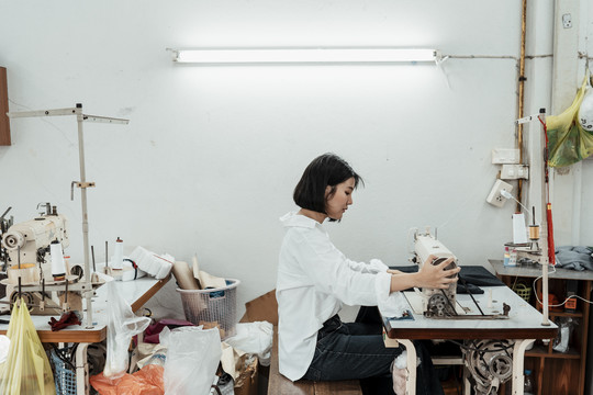 年轻的泰国女设计师坐在工作台上用黑布缝纫机。
