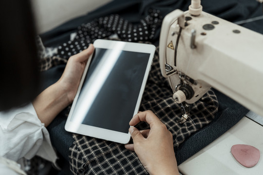 年轻的泰国女设计师在缝纫机站用平板电脑寻找制作服装的灵感。