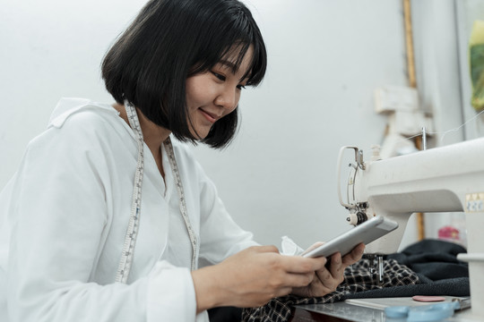 年轻的短发泰国女设计师在缝纫机站使用平板电脑。