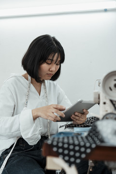 年轻的短发泰国女设计师在缝纫机站使用平板电脑。