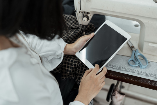 俯视图-身穿白衬衫的泰国女设计师在缝纫机桌旁使用平板电脑。