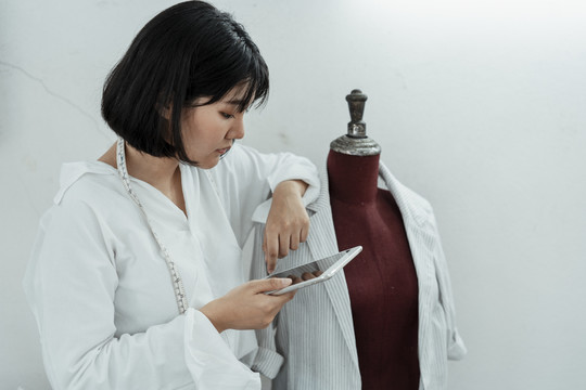 年轻的亚洲设计师正在考虑下一件衣服该做什么。手持平板电脑在互联网上搜索信息。
