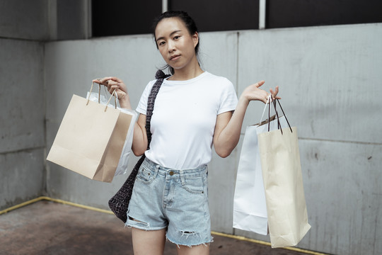 年轻的泰国亚裔女子，穿着白色衬衫和牛仔裤短裤，背着许多购物袋。