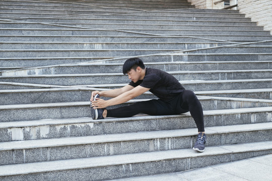 穿黑色运动服的运动型男子在城市锻炼前，会在楼梯上伸展双腿。