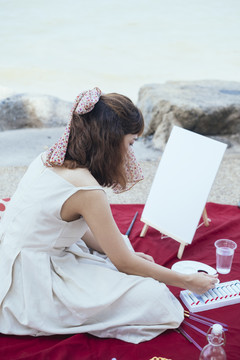 美丽的亚泰艺术家，穿着白色连衣裙，在夏天的海滩上，在画布上绘画之前，在调色板上混合颜色。