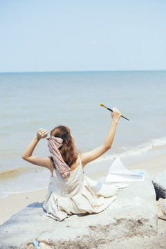 后视图-美丽的亚泰艺术家在白色礼服举行小刷子坐在沙滩上。