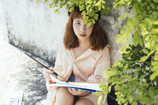 美丽的新潮亚泰艺术家穿着长衫，坐在树下用水笔在纸上作画。