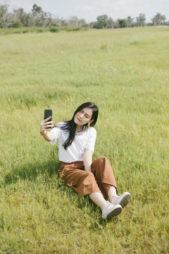 穿着休闲服的黑色长发女子坐在草地上，用智能手机自拍。