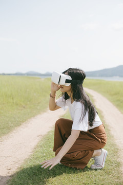 黑色长发女子穿着休闲服，戴着VR护目镜，在泥泞的草原上行走，享受虚拟现实技术。