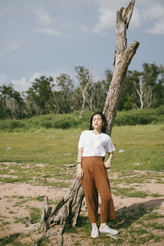 穿着休闲服的黑色长发女子站在土路上，靠在干枯的树上然后举手防晒。