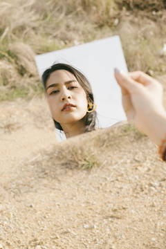 站在干燥的草原上，戴着耳环的白裙黑辫子女人在方镜下的倒影。