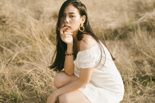 黑发女子身穿白裙，戴着耳环坐在干燥的草原上。