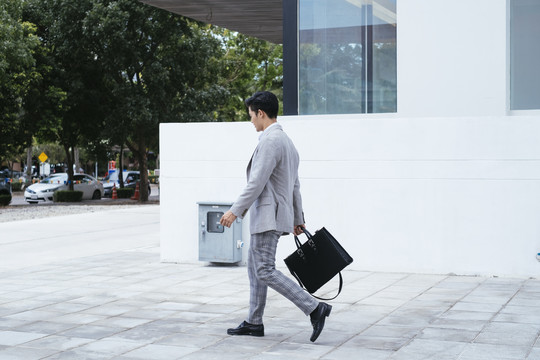 侧视图-聪明的亚泰商人穿着正装，手里拿着黑包走在街上。