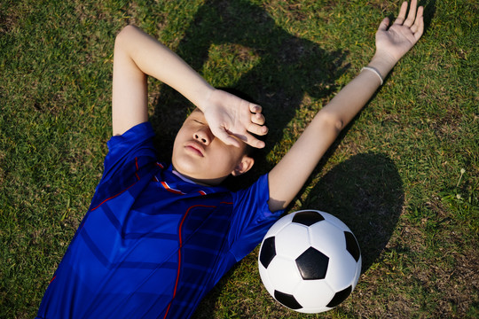 一个穿足球服的男孩躺在球场上，边睡边拿球。