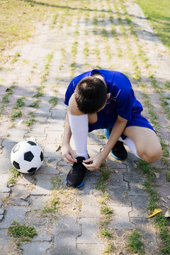 一个穿着蓝色足球服、系着鞋带的男孩站在球场旁边的人行道上。