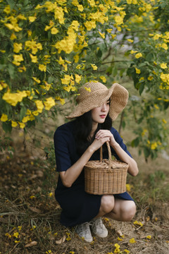 可爱的泰国女孩坐在黄花丛中，戴着帽子，拿着篮子。