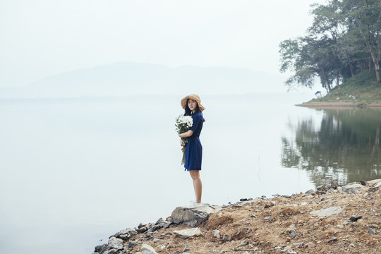 一个美丽的泰国女孩站在湖边的岩石上，怀里抱着白花，看着镜头。