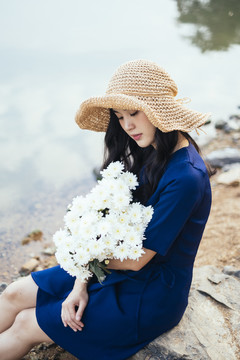 一个穿着深蓝色裙子的可爱女孩坐在湖边，爱上了怀中的白花。
