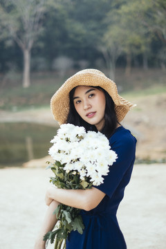 身着深蓝色连衣裙的可爱女孩抱着白花站在湖边，看着镜头。