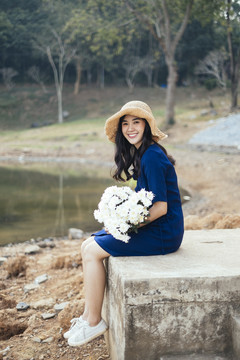 一个穿着深蓝色衣服的漂亮女孩，怀里抱着一朵白花，坐在湖边的岩石上。