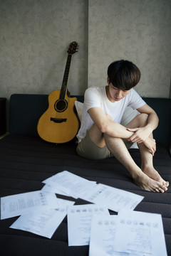 年轻的泰国吉他手坐在床上，拿着原声吉他，看着歌词笔记纸。