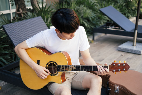 年轻的泰国吉他手在户外沙滩椅上弹原声吉他。旅行者吉他手。