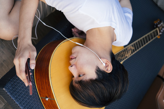 年轻的泰国吉他手躺在吉他上用耳机听智能手机里的音乐。