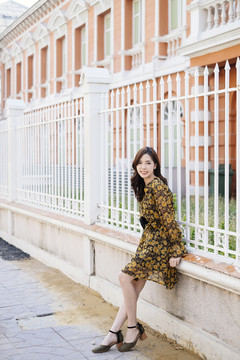一头卷曲的长发，穿着黄色连衣裙的女孩坐在大楼的栅栏边微笑着。
