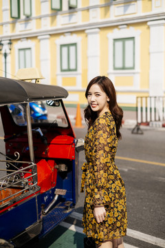 可爱的卷曲长发女孩穿着黄色连衣裙站在图克汽车旁边的道路前的粉彩建筑。