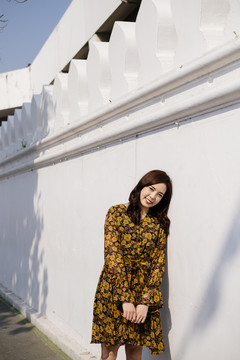 可爱的卷发女孩穿着黄色的裙子站在白色的墙上，在阳光下把头靠在墙上。