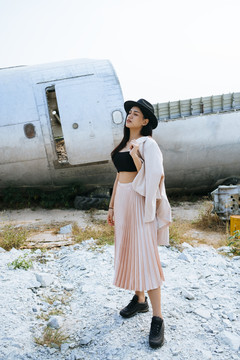 黑色长发女孩穿着华丽的衣服和粉色西装，戴着黑色的帽子站在废弃地区的废墟飞机上。