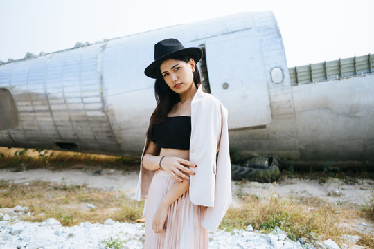 黑色长发女孩穿着华丽的衣服和粉色西装，戴着黑色的帽子站在废弃地区的废墟飞机上。