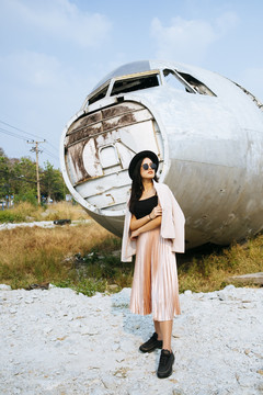 黑色长发女孩穿着粉色西装，戴着黑色帽子和墨镜站在废弃地区的废墟飞机前。