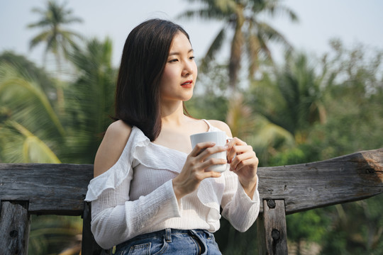 一位泰国亚裔妇女坐在乡村地区的木凳上喝咖啡。在大自然中旅行。在一杯咖啡中加香。喝白咖啡的漂亮女人。