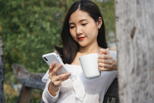 一位身穿白衬衫的亚泰妇女端着一杯咖啡，坐在木凳上，用智能手机与朋友联系。