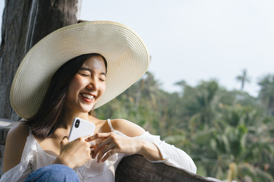 一位兴高采烈的亚洲妇女手持智能手机戴着帽子坐在阳台上。