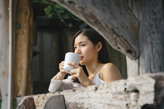 在乡村旅馆的木制阳台上，一名亚泰妇女拿着一杯咖啡向外眺望。